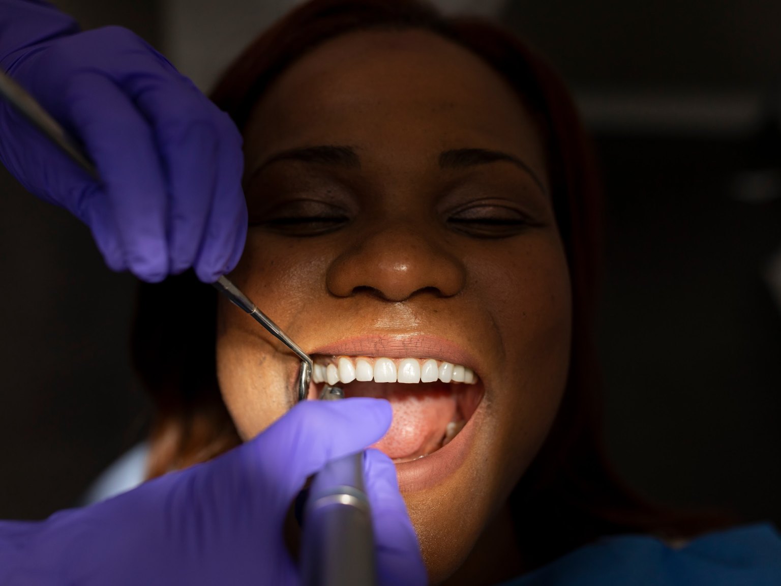 A patient smiling with eyes closed while gloved hands hold a mirror and handpiece at her teeth