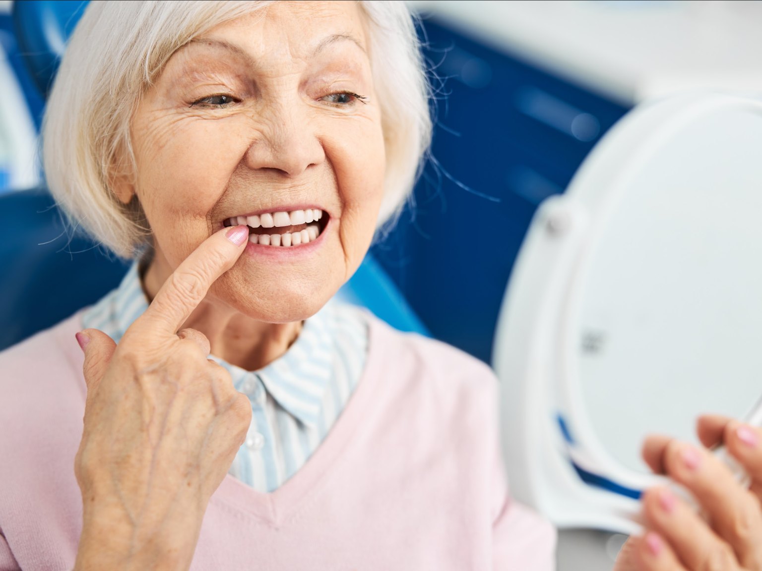 An older woman pointing at her new teeth while checking them in a hand mirror