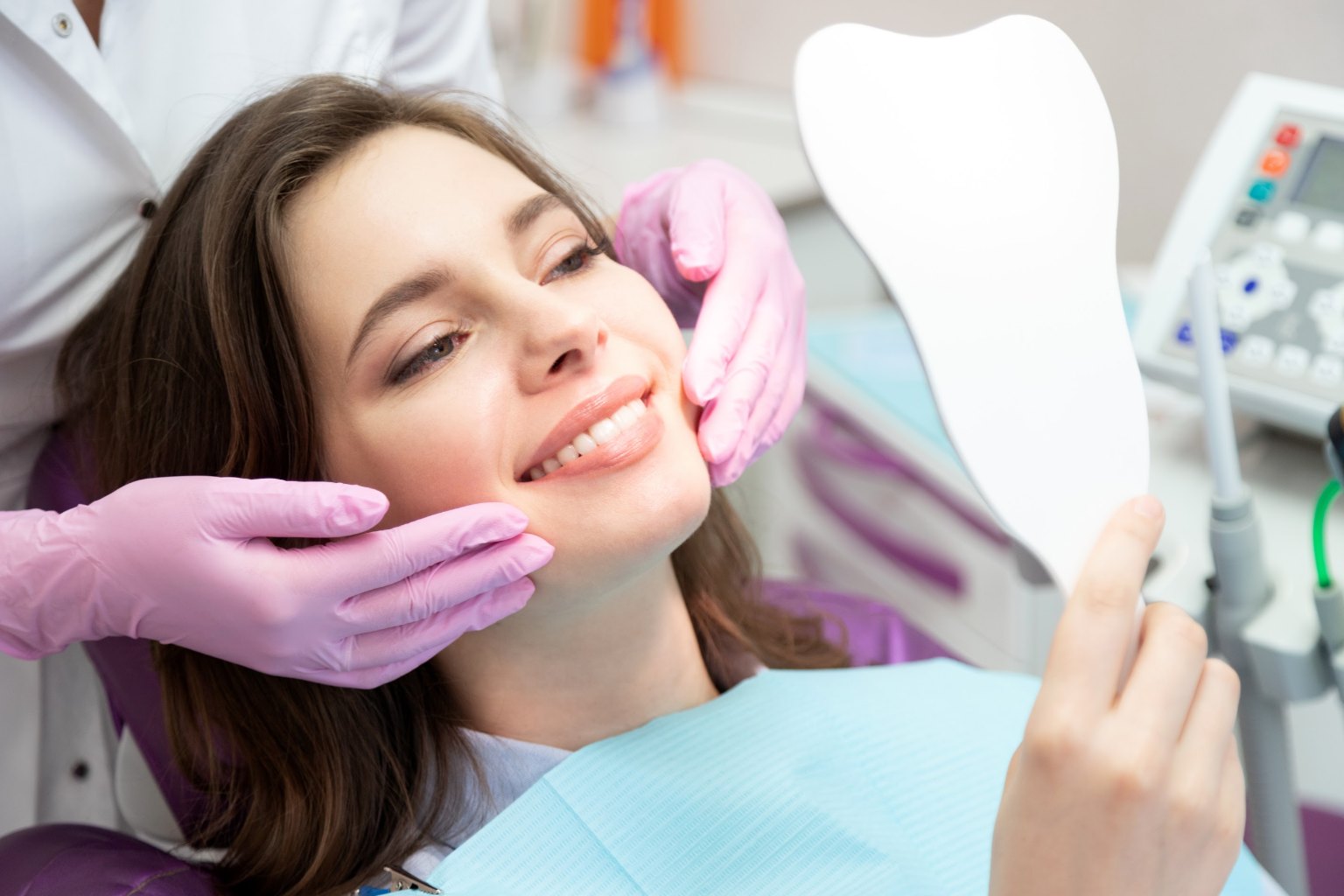A patient smiling at her reflection in a hand mirror as the dentist gently holds her jaw