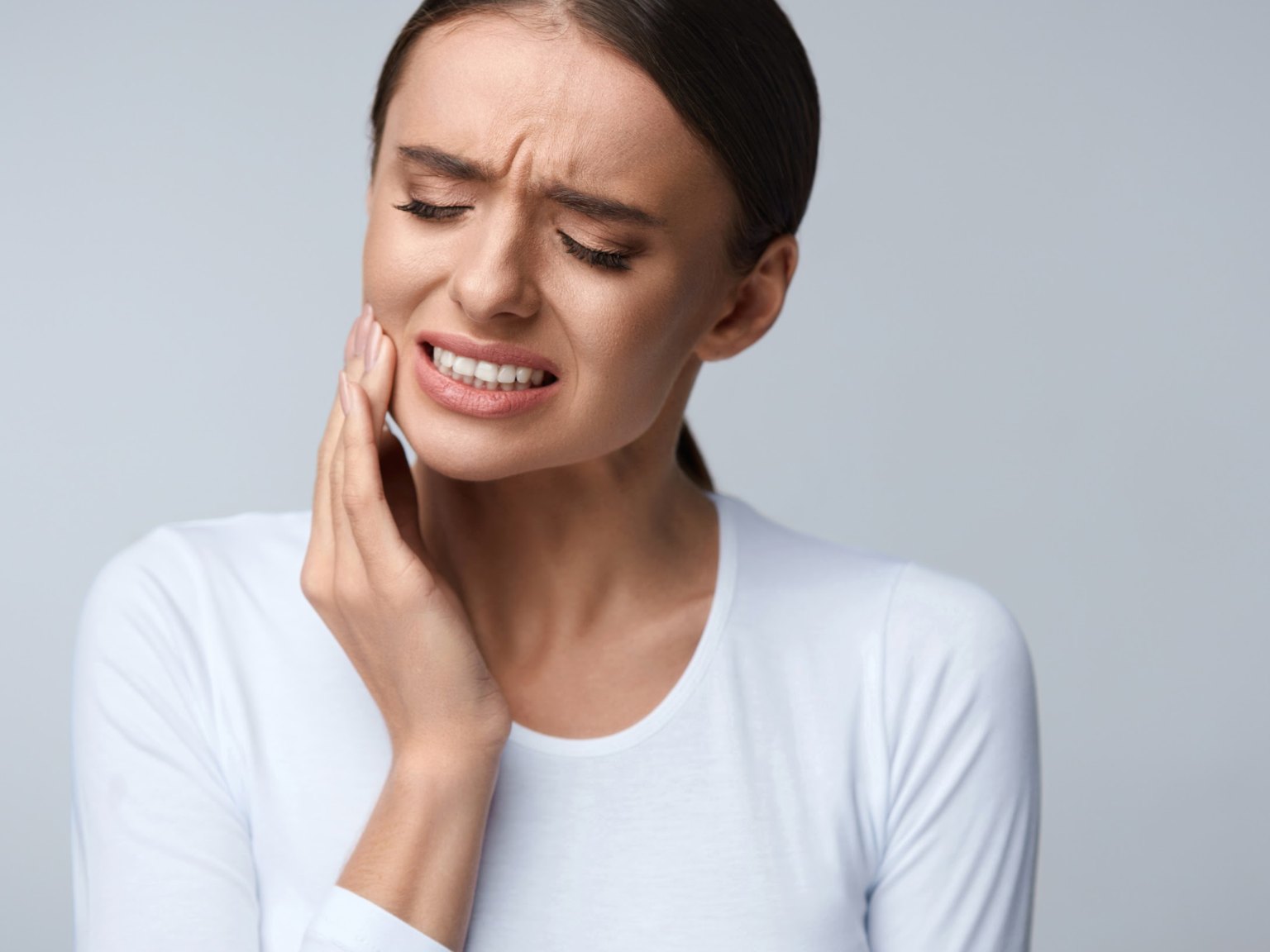 A woman wincing with her eyes shut, holding her cheek over an aching tooth