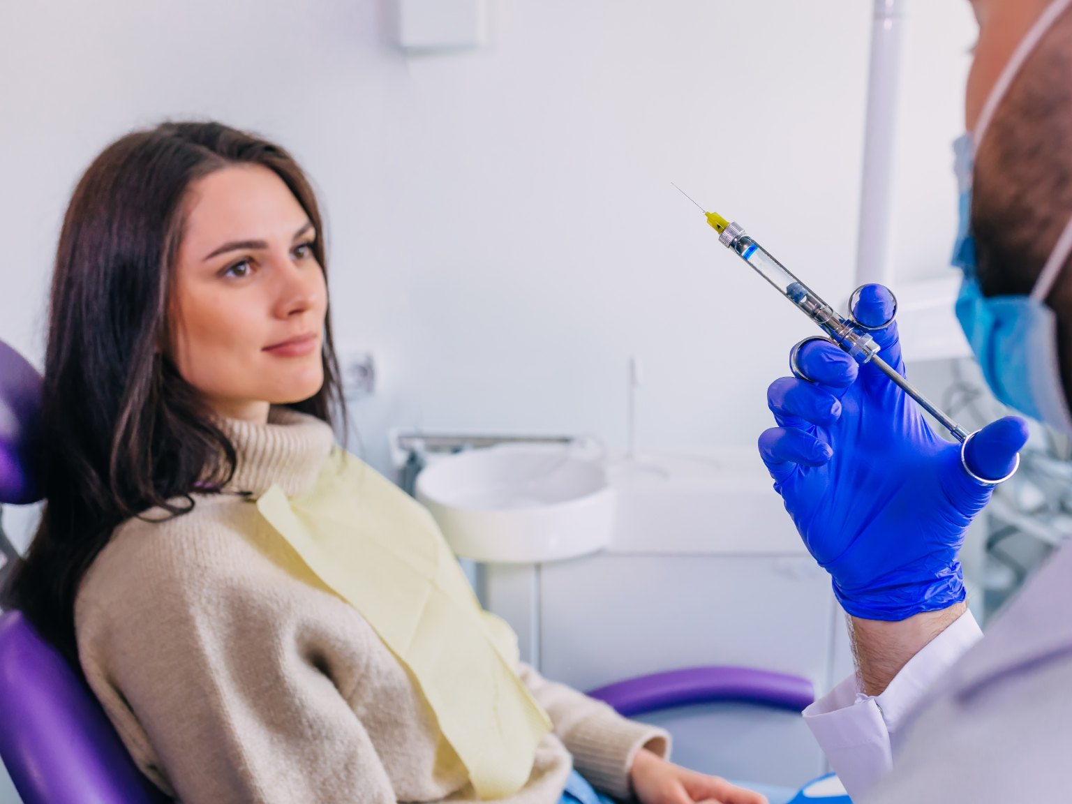 A woman in the dental chair watching a gloved hand hold up an anaesthetic syringe