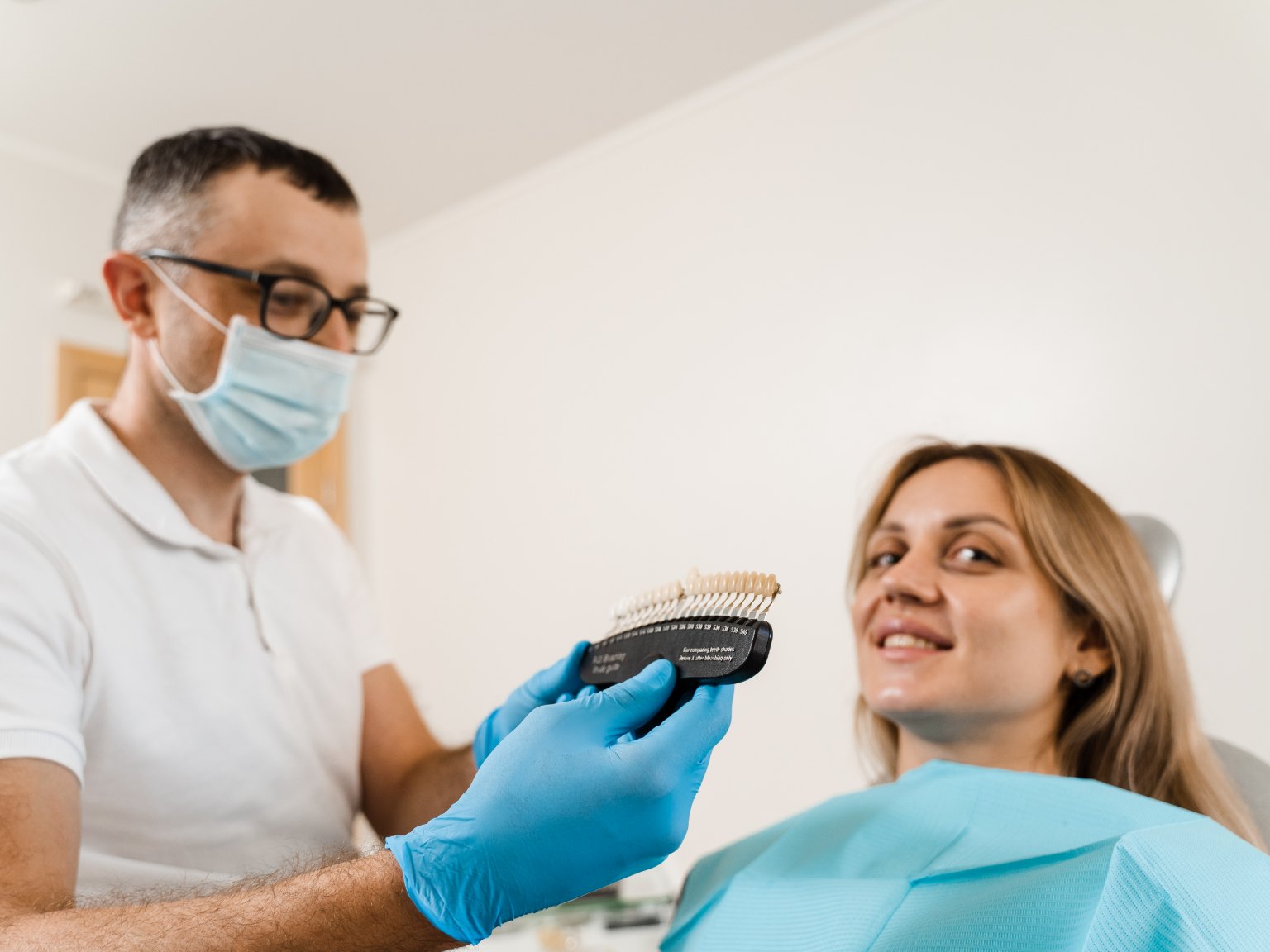 A dentist holding a tooth shade guide up to a smiling patient to match a new crown