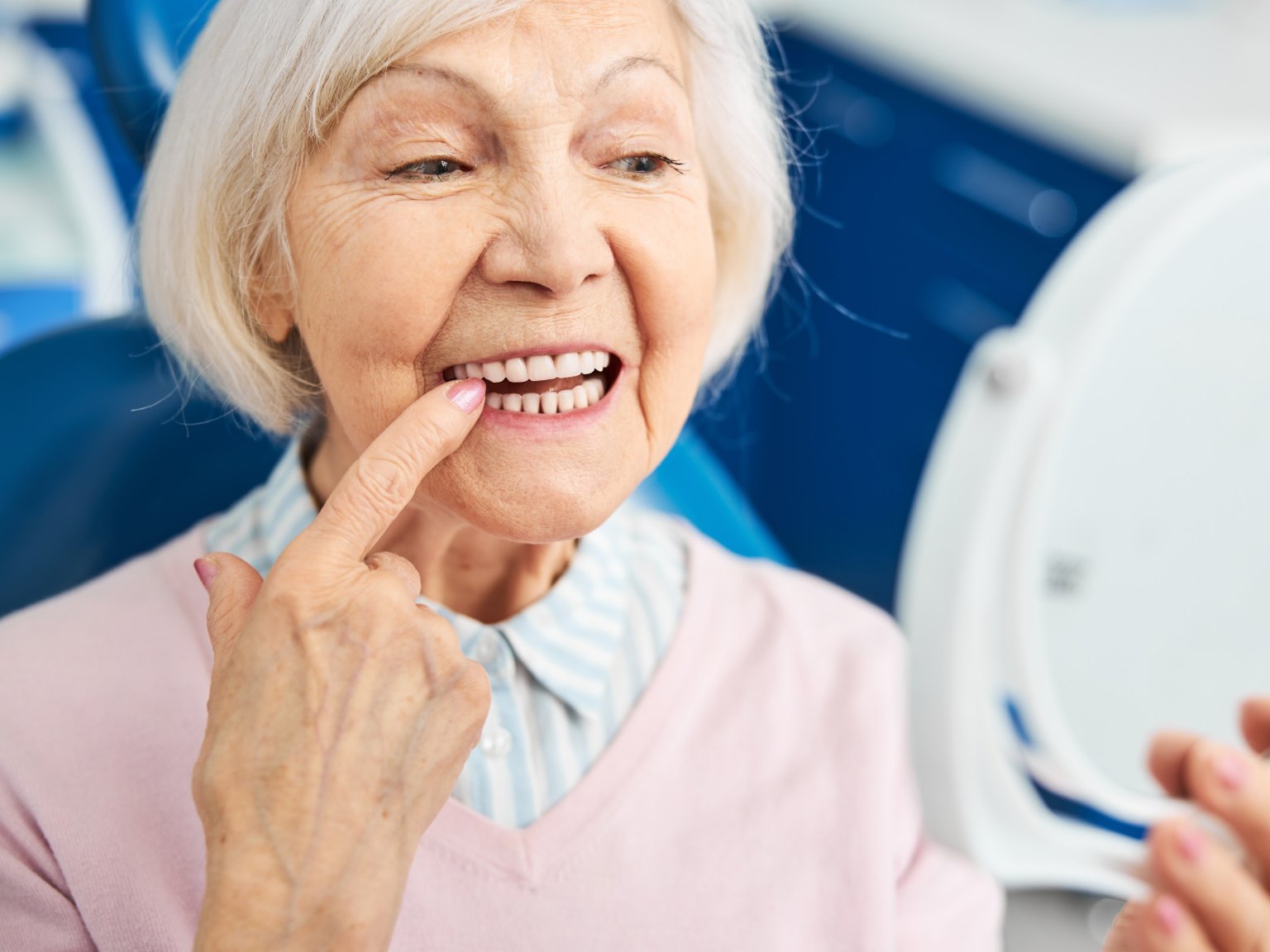 An older woman in a dental chair checking her gums and teeth in a hand mirror