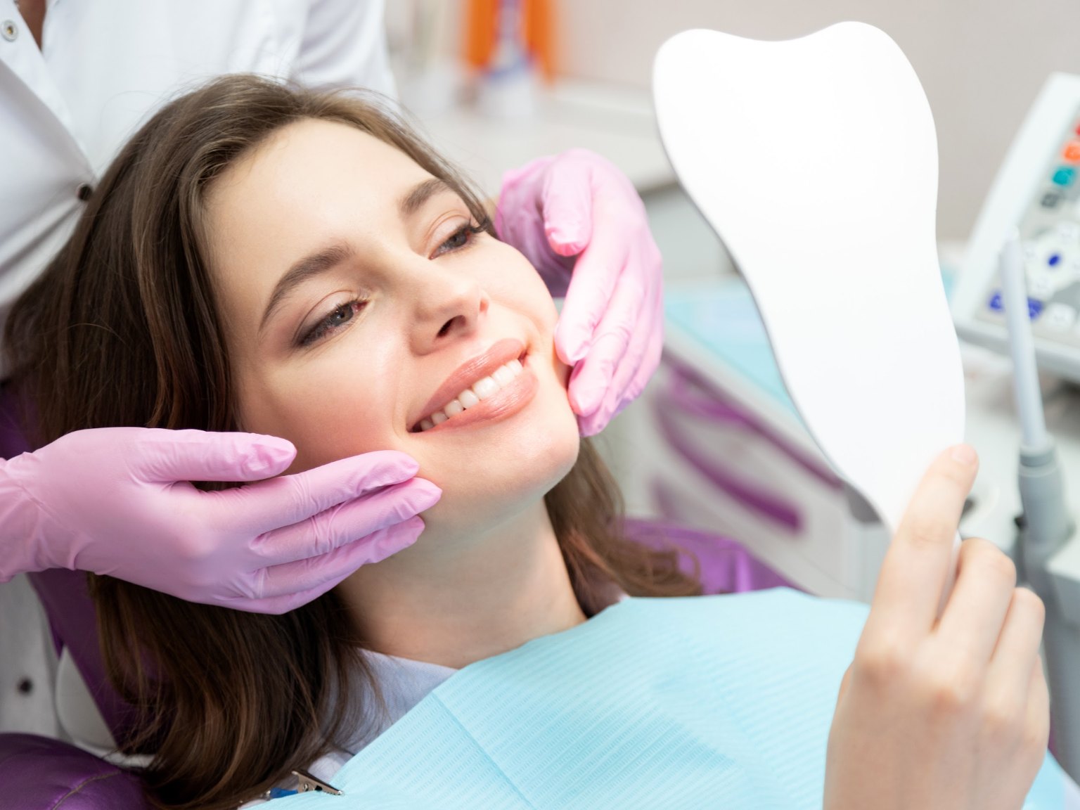 A patient in the chair smiling at her teeth in a hand mirror as gloved hands frame her face