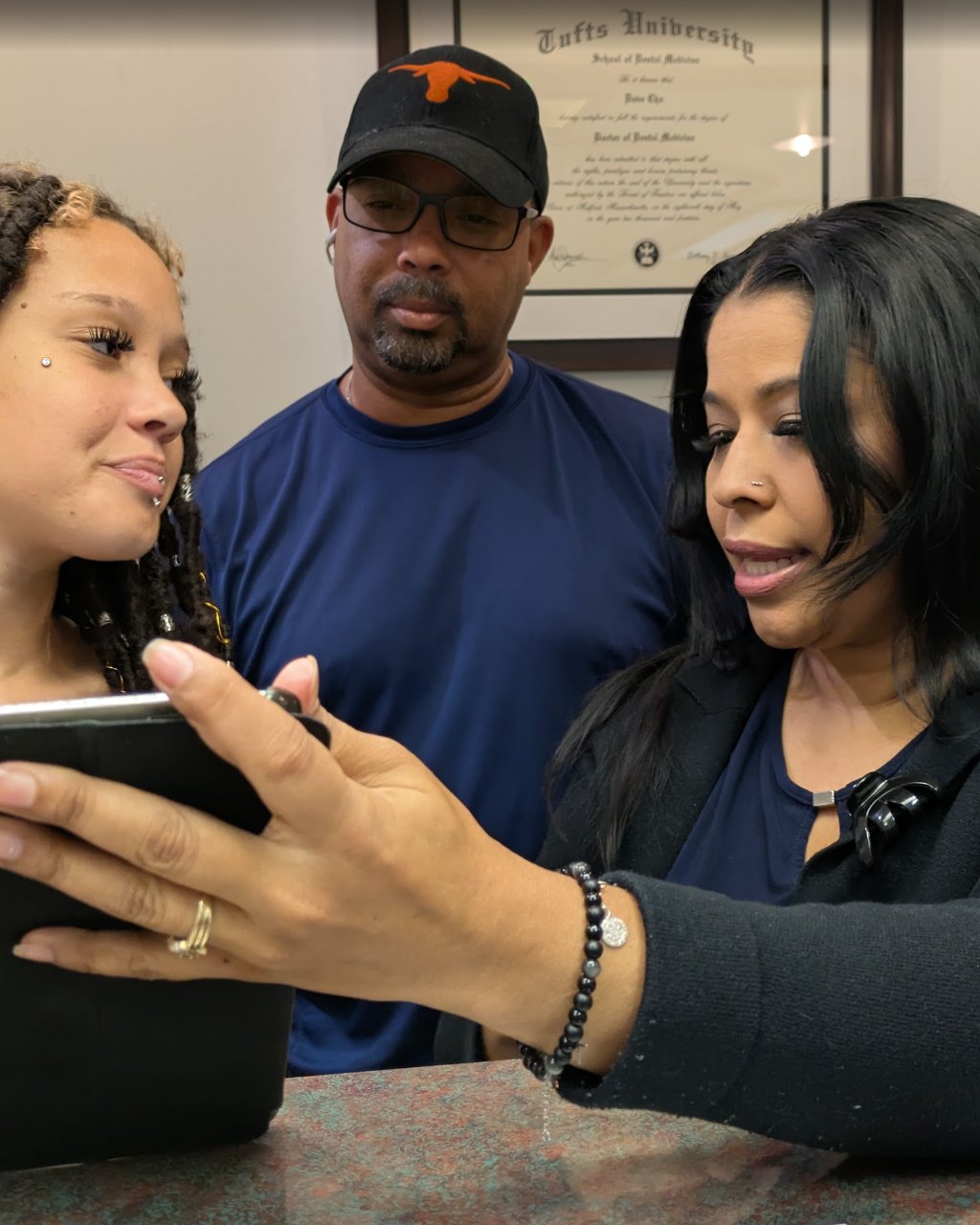 A team member showing two patients a tablet at the front desk, a diploma behind them