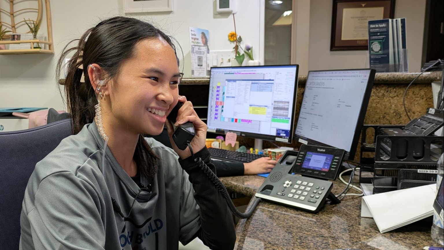 A receptionist smiling on the phone at the front desk, monitors and phone beside her