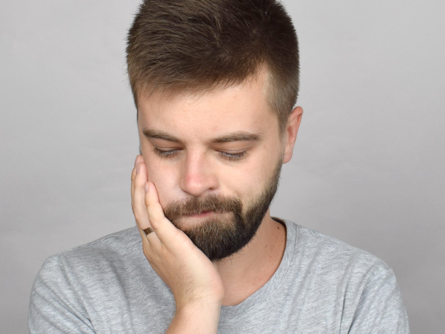 A young bearded man looking down with a hand pressed to his back jaw